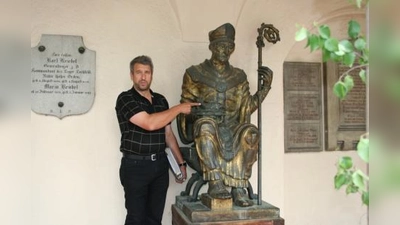 Der Namenspatron der Kirche St. Ulrich empfängt die Besucher des Gotteshauses im Kreuzgang. Der Hobby-Historiker Norbert Winkler deutet auf die Symbole des Heiligen – Fisch und Evangeliar. (Foto: tg)