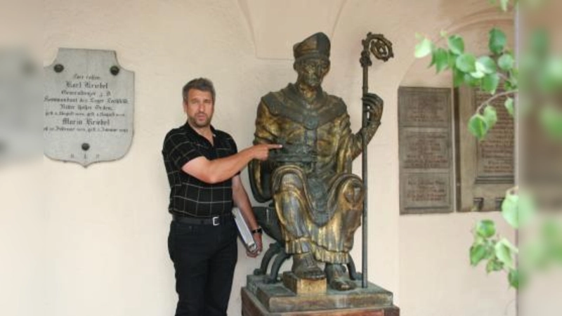 Der Namenspatron der Kirche St. Ulrich empfängt die Besucher des Gotteshauses im Kreuzgang. Der Hobby-Historiker Norbert Winkler deutet auf die Symbole des Heiligen – Fisch und Evangeliar. (Foto: tg)