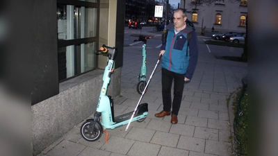 Am Laimer Platz erklärt Stefan Unterstraßer, warum auf dem Gehweg zurückgelassene Roller für ihn gefährliche Stolperfallen sind. Zudem machen es ihm die Scooter wie hier unmöglich, sich mit seinem Taststock an der Kante entlang des U-Bahn-Aufzugs und des Gehweg zu orientieren. (Foto: job)